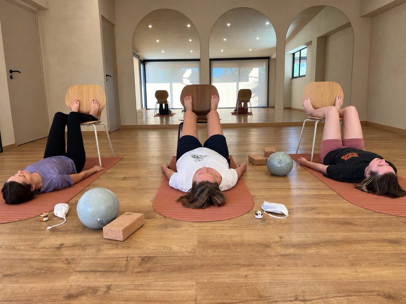 Trois élèves allongées sur leur tapis, jambes posées sur une chaise, dans une salle lumineuse aux arches blanches